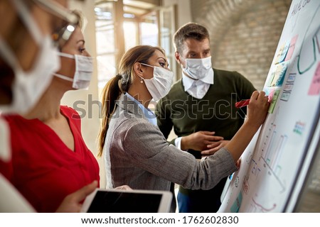 Young businesswoman wearing protective face mask while writing mind map on whiteboard and making new business plans with her team during COVID-19 pandemic. 