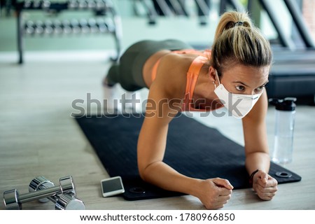 Female athlete exercising strength in plank pose while wearing protective face mask at health club. 