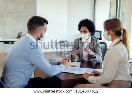 Black real estate agent and a couple wearing protective face masks while communicating during the meeting in the office. 
