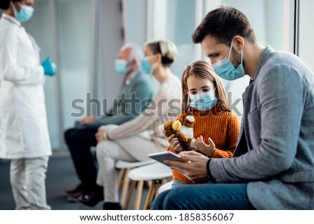Father and daughter wearing protective face masks while surfing the net on digital tablet in a hallway at the hospital.  Imagine de stoc © 