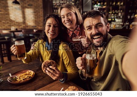 Carefree friends taking selfie while drinking beer and eating hamburgers in a bar. 