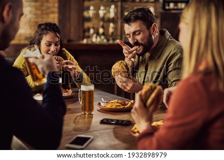 Group of friends drinking beer and eating hamburgers in a pub. Focus is on man tasting a burger. 