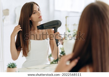 Beautiful girl using a hair dryer and smiling while looking at the mirror. Smiling woman drying hair with hair dry machine. Happy girl looking at mirror while using hair dryer in the bathroom 