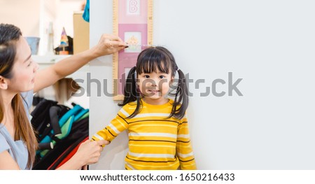 Mother Height measure her daughter near white wall at home.2.6 years old girl kid standing and looking at above using hand measuring her height.Growth hormones, child development, Calcium concept.