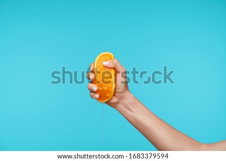 Indoor photo of young attractive hand holding orange and clenching a fists while queezing juice, preparing breakfast while posing over blue background Stockfoto © 