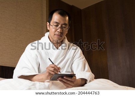 Mature Asian man in bathrobe in hotel, writing note.
