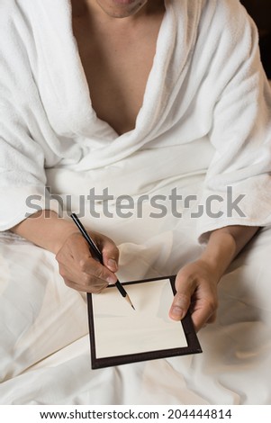 Mature Asian man in bathrobe in hotel, writing note.
