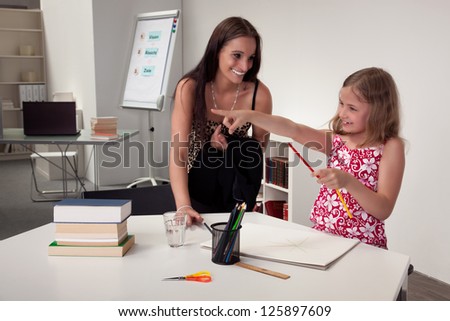 Enthusiastic attractive young girl in art class with her teacher or mother pointing her hand and smiling, interior portrait in a classroom or office
