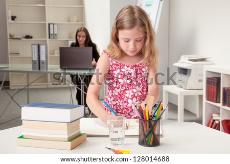 Pretty little girl sketching at a table in a classroom or home office with a teacher or her mother sitting in the background working at a laptop and keeping an eye on her