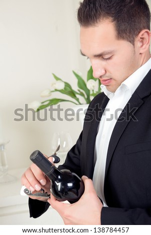 Stylish handsome young man in evening wear standing opening a bottle of red wine that he is holding in his hand
