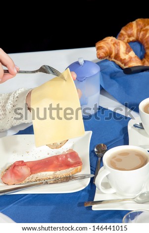 Woman seated at a breakfast table helping herself to sliced cheese to add to her wholewheat roll with ham