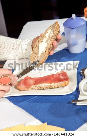 Hands of a woman seated at a table buttering a roll for breakfast with one half already on her plate topped with cold meat
