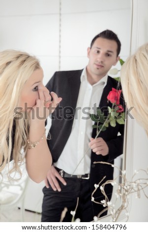 Romantic man carrying a single long stemmed red rose reflected in a mirror admiring and watching his sweetheart is she titivates in the mirror