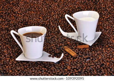 A cup of coffee and a cup of milk, placed on spilled coffee beans and cinnamon