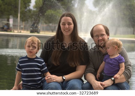 Family of 4, including mother, father, preschool boy and toddler daughter, with a fountain behind them