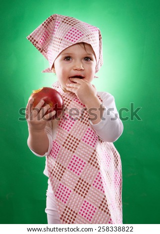 the baby girl with a kerchief and kitchen apron holding an vegetable isolated on the green background