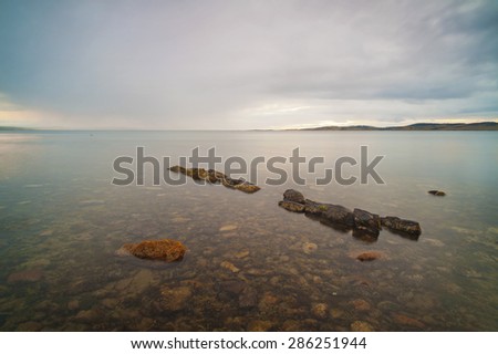 The rocky ocean floor can be seen through the very clear water, broken only by a small line of rocks pointing to the horizon.