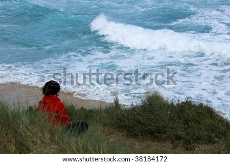 A woman in a red top sits on top of a sand dune looking out over the ocean