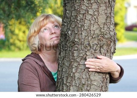 A natural mature woman looking upwards with an arm around tree in a suburban neighborhood.