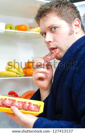 Handsome man eating ham slice near open fridge