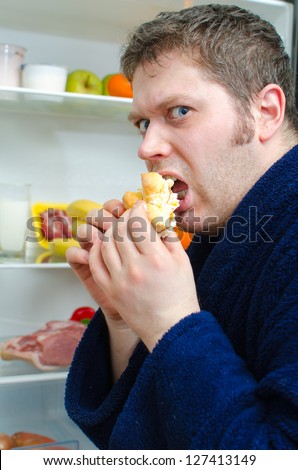 Handsome man eating piece of cake near open fridge