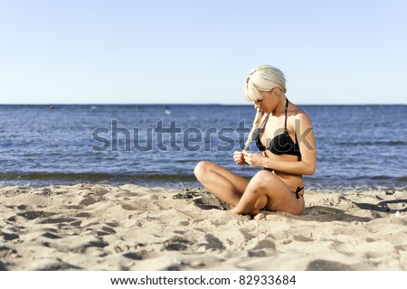 blonde girl in a black bathing suit sitting on shore blue sea on the sand