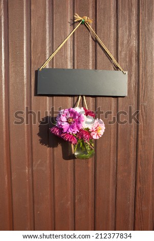 A rustic hanging decoration made from a jam-jar full of summer flowers beneath a blank sign suspended from string on a wooden board background