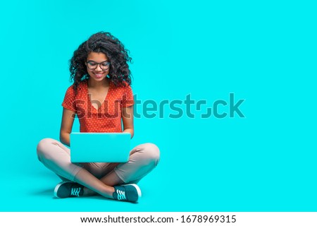 Beautiful young smiling woman in casual outfit and trendy eyeglasses sitting isolated on bright colored blue background and working on her laptop. Full length horizontal studio shot. Copy space.