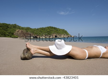 beautiful woman in white bikini lying on sand on tropical beach with feet resting on coconut and a white hat on her knees