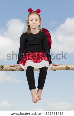 little girl in lady bug costume seated on a branch against a blue sky