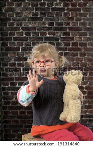 little toddler with glasses and teddy bear saying hello against a brick wall