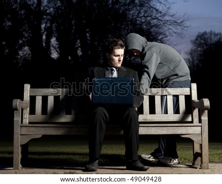 Business man sitting on a park bench, using his laptop, whilst a hooded stranger attempts pickpocket him.