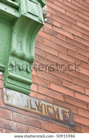 An old rusted street sign on a red brick wall, with the corner of a green window casing, that looks to have been painted over and over again.