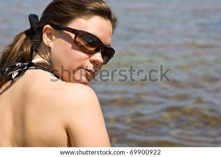 A woman with her back to the camera, her bare back and bathing suit strap visible, looking back towards the camera, with water in the background.