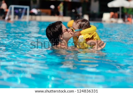 In the sunny day in the summer the boy in sun glasses and in a life jacket plays pool with mum