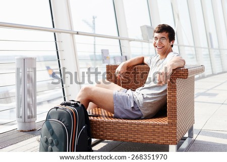 Happy young man in glasses and headphones sitting on chair in airport departure lounge