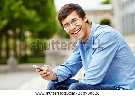 Portrait of smiling young man in glasses and blue shirt sitting in park and using mobile phone