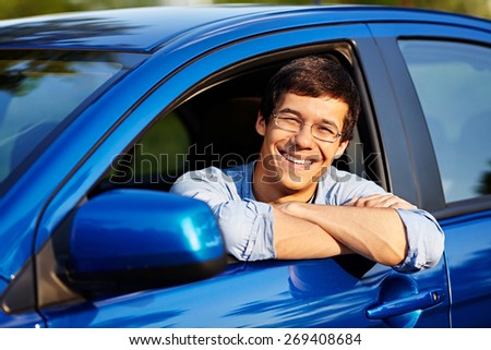 Portrait of young handsome smiling man in glasses looking out through open car window outdoor in sunny day