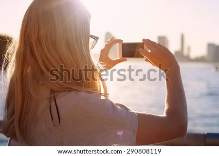 Closeup of blonde girl in glasses taking picture of city bay with mobile phone on sunset. Focus on glasses