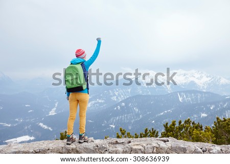 Back view of young woman wearing pink hat, blue jacket, green backpack, yellow pants and hiking boots raising her hand against winter mountains celebrating successful climb - goal concept
