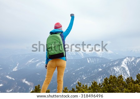 Back view of young woman wearing pink hat, blue jacket, green backpack and yellow pants raising her hand against winter mountains celebrating successful climb - goal concept