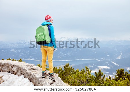 Back view of young woman wearing pink hat, blue jacket, green backpack, yellow pants and hiking boots standing against winter mountain valley - adventure concept