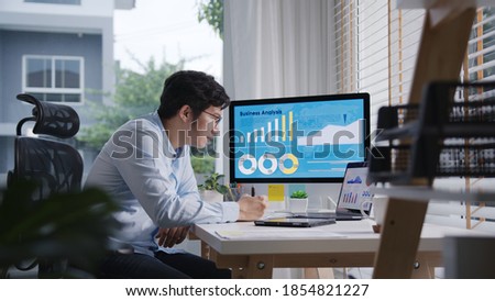Candid of young attractive asian thai man busy work multiple screen computer or smart tablet on table desk at home in freelance data analyst, data science scientist for business.