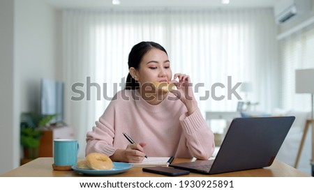 Young attractive beautiful asian female hungry eat doughnut take away snack food with full mouth look at computer notebook at home in busy work from home multitask unhealthy meal lifestyle concept.