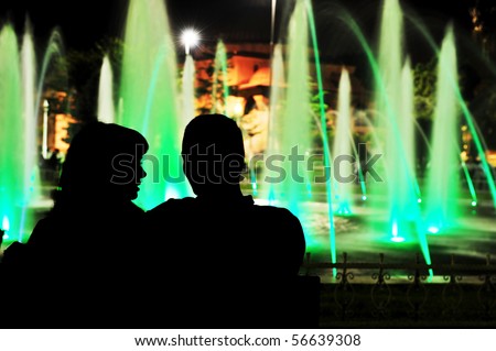 Beautiful night scene of young couple in light park, lovers silhouettes  against green fountain