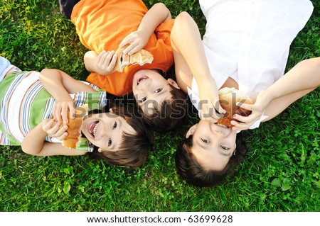 Three children laying on green grass on ground and eating sandwiches and smiling, healthy food, good friendship and love and happiness without limit :)