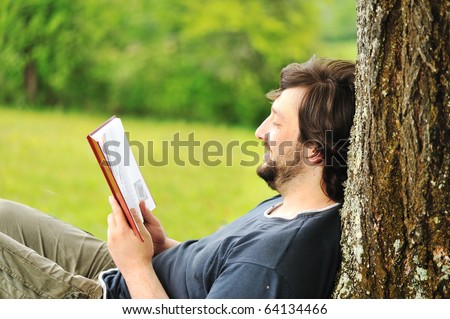 Young relaxed man reading book in nature, back on tree, meadow behind
