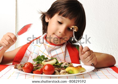 Cute positive boy with fork and knife eating on lunch table