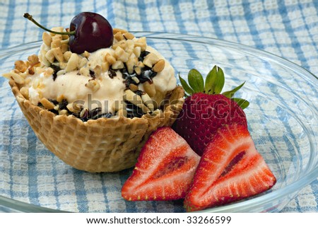 Vanilla Ice Cream Sundae in an edible waffle bowl with chocolate syrup, nuts, strawberries on the side, and a cherry on top sitting on clear plate on blue and white checked tablecloth