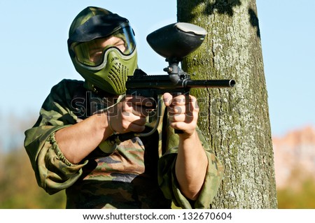 paintball player in protective uniform and mask aiming and shooting with paint marker gun outdoors